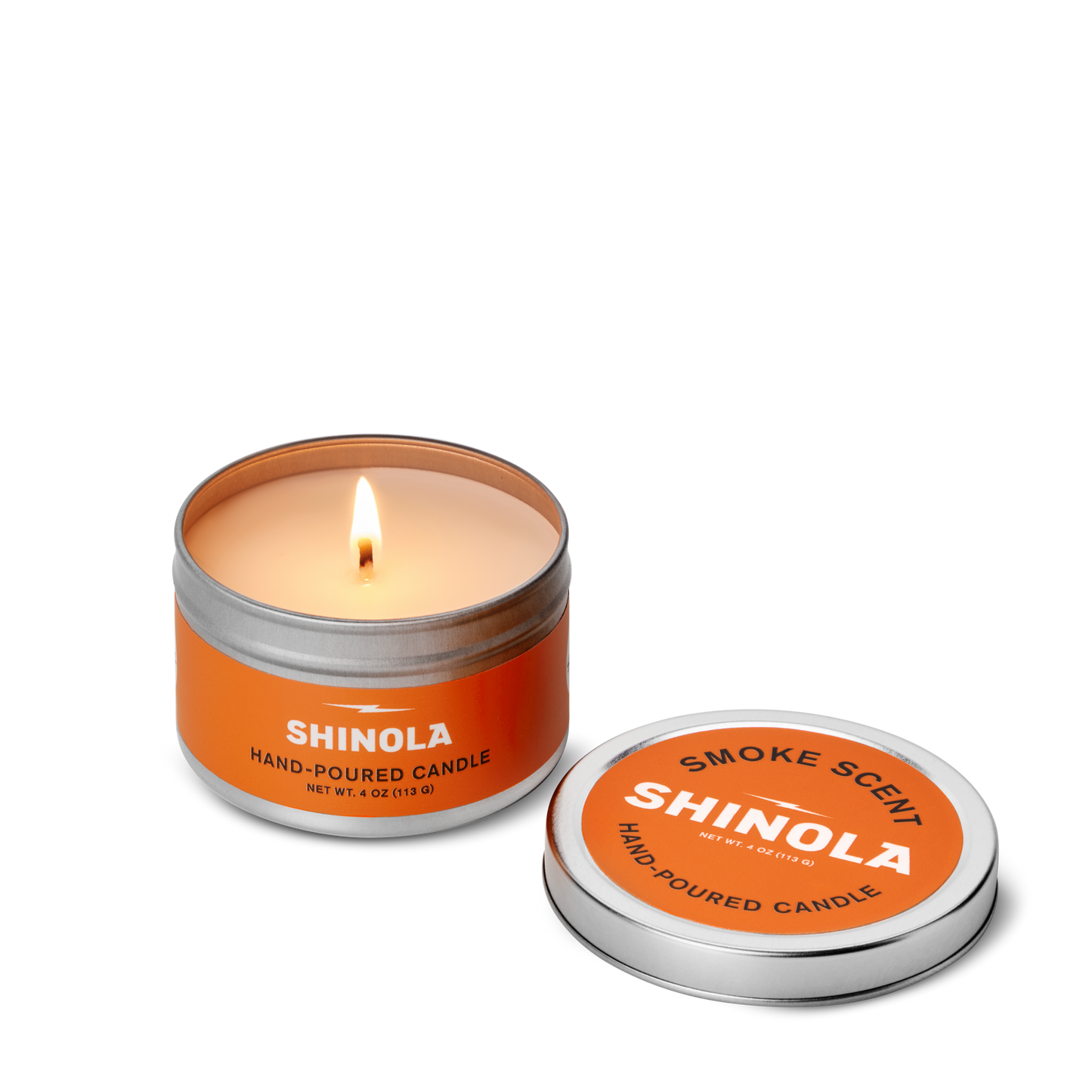 A lit Shinola Hand Poured 4oz Travel Candle - Smoke rests in an orange tin labeled SHINOLA, crafted in Detroit, with a removable lid beside it displaying "Smoke Scent SHINOLA" on a white background.