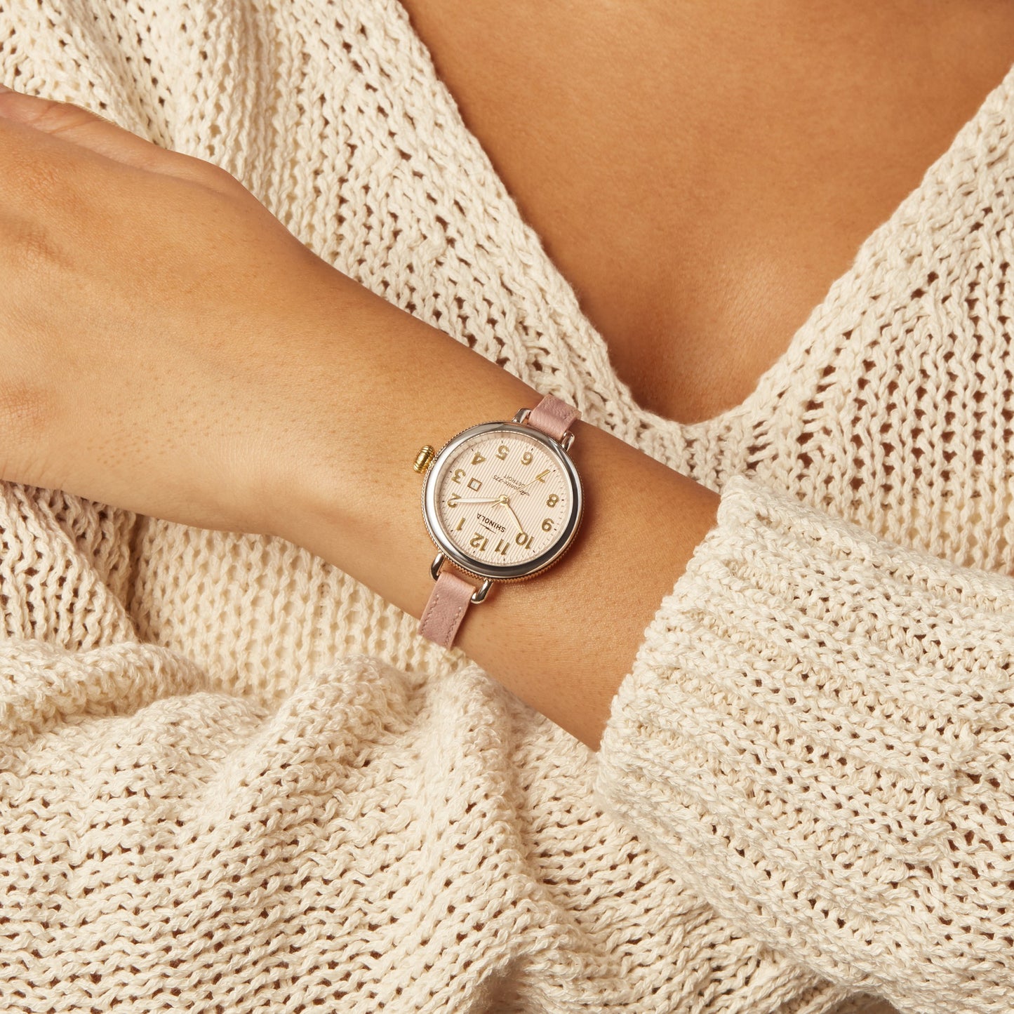 A person in a cream knit sweater showcases the Shinola Birdy Watch - Light Pink, featuring a pink dial with gold numbers and hands, gold accents, and a light pink leather strap on their left wrist.